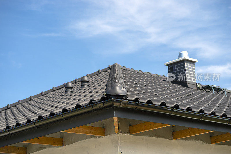 The roof of a single-family house covered with a new ceramic tile in anthracite against the blue sky, visible ridge tile, system chimney and ceramic ventilation fireplace on the roof.